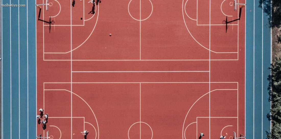 An aerial view of a large, vibrant sports complex featuring a red basketball court with multiple hoops and a surrounding blue running track. Several people are scattered across the area, engaged in various activities.