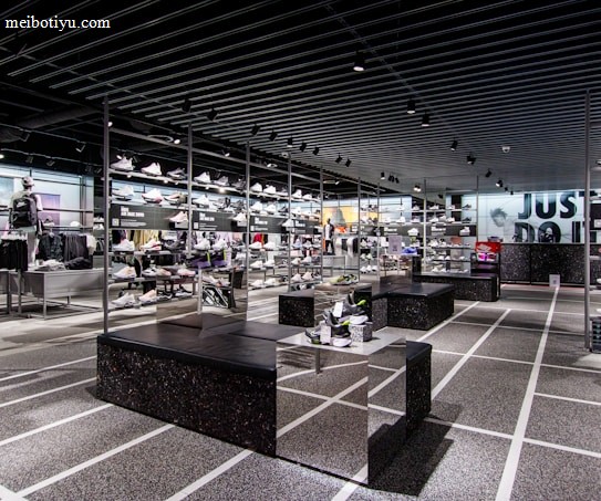 A modern sneaker store with sleek black shelving displaying various athletic shoes and sports apparel. Central seating areas and spotlights provide a contemporary retail ambiance. The signage prominently features a famous sportswear slogan in the background.