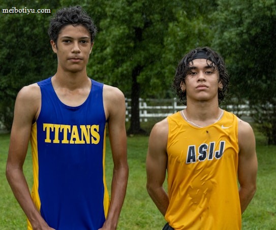 Two young athletes wearing sleeveless sports tops stand on a grassy field. The athlete on the left is wearing a blue top with the word 'TITANS' while the one on the right wears a yellow top with 'ASIJ'. The background features green trees and a white fence, with a bench visible on the right.