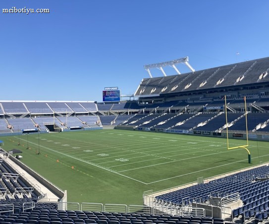 A large, empty sports stadium with blue seats surrounding a well-maintained green grass field. Bright sunlight creates clear shadows, and there are two goal posts on opposite ends of the field. A big electronic scoreboard displays some graphics, and the sky is clear and blue.